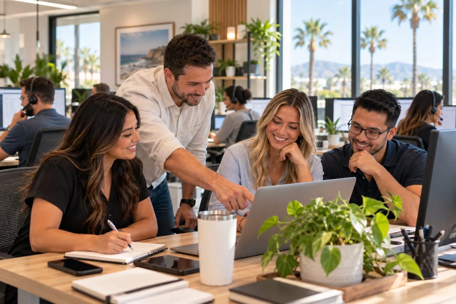 Small business team smiling around a laptop in a sunny office with palm trees outside and a help desk team in the background